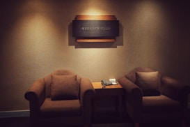 A cozy and dimly lit room features two comfortable brown armchairs positioned on either side of a small wooden table. Mounted on the wall above the table is a decorative sign that reads 'Regency Club.' The table holds a classic telephone and some note pads.