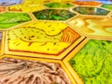 A close-up view of a board game with hexagonal tiles featuring different landscapes such as fields, forests, and deserts. The tiles exhibit vibrant colors and detailed textures with each hexagon bordered distinctly.