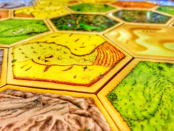 A close-up view of a board game with hexagonal tiles featuring different landscapes such as fields, forests, and deserts. The tiles exhibit vibrant colors and detailed textures with each hexagon bordered distinctly.