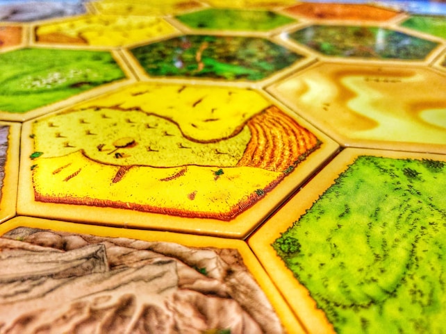 A close-up view of a board game with hexagonal tiles featuring different landscapes such as fields, forests, and deserts. The tiles exhibit vibrant colors and detailed textures with each hexagon bordered distinctly.