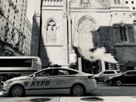 A New York City police traffic vehicle is parked on a street near a historic church with Gothic architecture. Smoke or steam rises from a vent on the sidewalk, and other vehicles, including a bus, are visible in the background. Tall buildings surround the scene, casting shadows on the street.