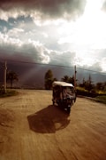 A motorized rickshaw is traveling on a dirt road, casting a long shadow in the late afternoon sun. The road is surrounded by lush greenery and palm trees with a mountain range in the distance. Dramatic clouds fill the sky, partially obscuring the sunlight.