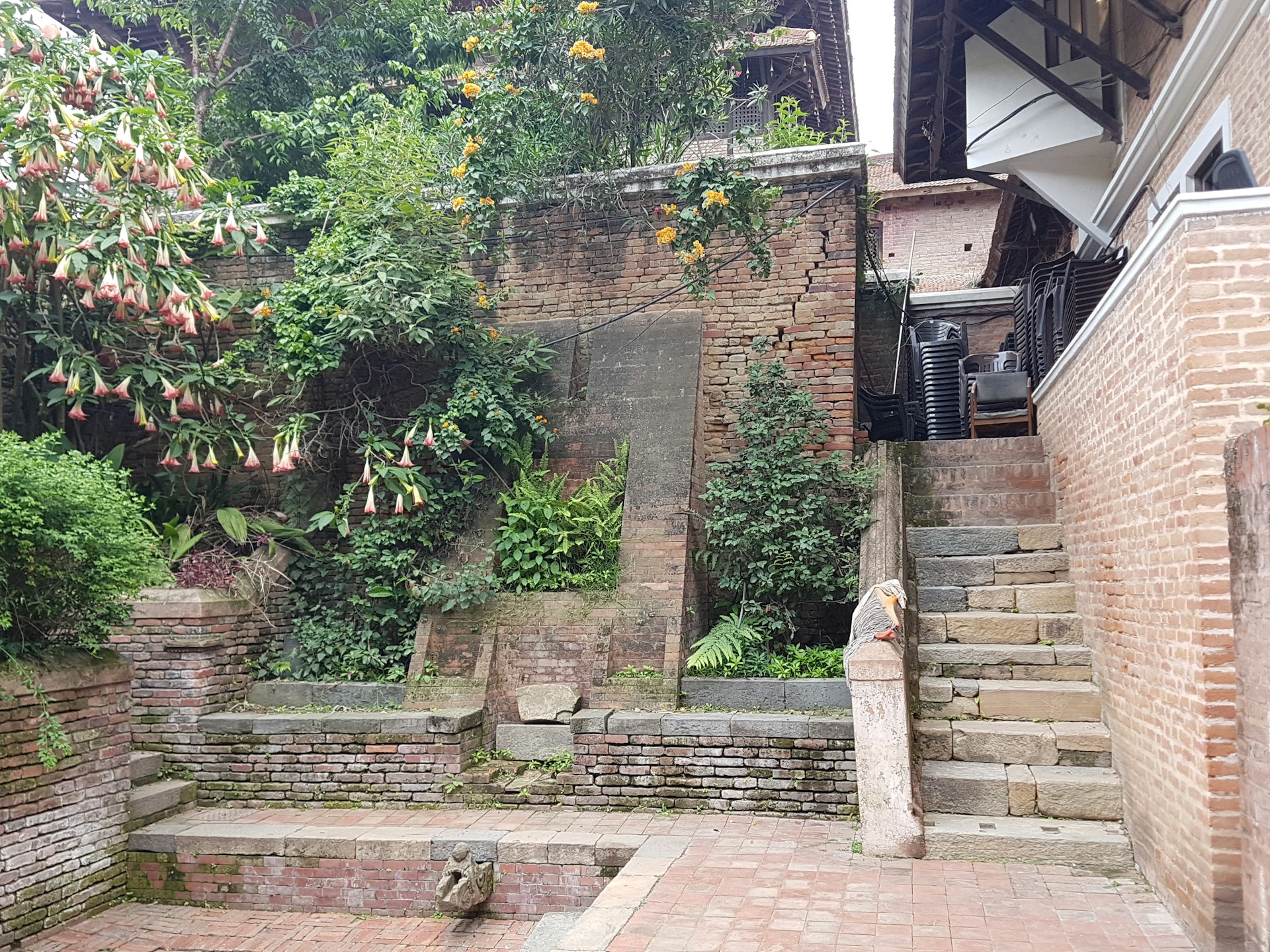A rustic courtyard featuring old, textured brick walls adorned with lush greenery and flowering plants. A stone staircase on the right leads upward, and the setting is complemented with stacked chairs and steps made from stone slabs.