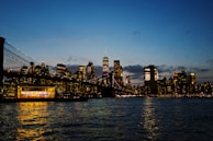 A city skyline at dusk with illuminated skyscrapers reflecting on a body of water. The Brooklyn Bridge is visible, and there is a brightly lit venue near the water's edge.