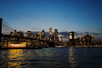 A city skyline at dusk with illuminated skyscrapers reflecting on a body of water. The Brooklyn Bridge is visible, and there is a brightly lit venue near the water's edge.