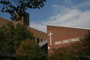 A brick building with a prominent cross and the text 'Freie Christengemeinde' on its facade is partially obscured by leafy trees. The structure's architectural design includes a tower and angular rooflines, set against a partly cloudy blue sky.