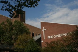 A brick building with a prominent cross and the text 'Freie Christengemeinde' on its facade is partially obscured by leafy trees. The structure's architectural design includes a tower and angular rooflines, set against a partly cloudy blue sky.