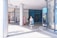 A woman is walking in an open-air shopping area carrying a blue shopping bag. She is dressed in a white shirt and a patterned skirt. Several large columns support the covered walkway. People can be seen in the background near a storefront with glass doors and windows. A person is sitting in a wheelchair nearby, and another is standing beside them.