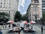 Two hot dog stands are situated on a city street, under red and white umbrellas with signage for cold drinks and hot dogs. People are casually walking around, and the area has a busy urban vibe. Classic tall buildings line the street, with lush green trees adding a touch of nature.