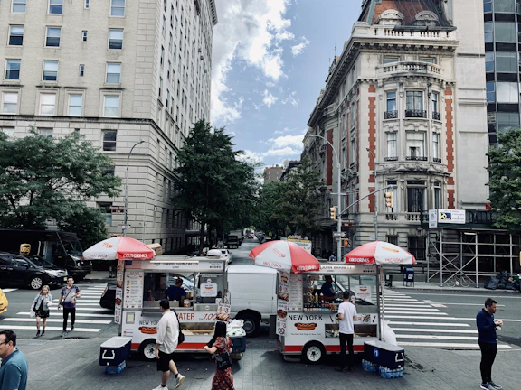 Two hot dog stands are situated on a city street, under red and white umbrellas with signage for cold drinks and hot dogs. People are casually walking around, and the area has a busy urban vibe. Classic tall buildings line the street, with lush green trees adding a touch of nature.