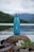 A blue water bottle with a silver cap is placed on a rock by a calm lake. The background features a scenic view of forested hills and misty mountains under a cloudy sky. There are some purple and white flowers around the rock, adding a natural element to the scene.