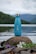 A blue water bottle with a silver cap is placed on a rock by a calm lake. The background features a scenic view of forested hills and misty mountains under a cloudy sky. There are some purple and white flowers around the rock, adding a natural element to the scene.