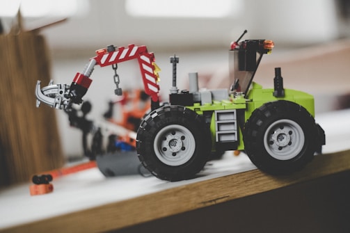 A toy tractor made from colorful interlocking plastic blocks, featuring black wheels and a green body. It has a movable arm ending in a claw, which is predominantly red and white.