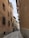 A narrow cobblestone street flanked by tall, closely packed buildings with beige and brown facades. The buildings have wooden window shutters and metal grilles. A sign is visible, indicating property for rent, and a person stands in the street looking at a phone.