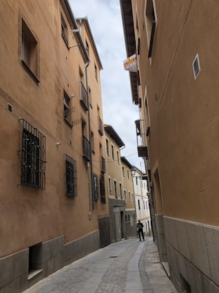 A narrow cobblestone street flanked by tall, closely packed buildings with beige and brown facades. The buildings have wooden window shutters and metal grilles. A sign is visible, indicating property for rent, and a person stands in the street looking at a phone.