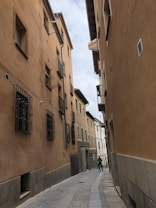 A narrow cobblestone street flanked by tall, closely packed buildings with beige and brown facades. The buildings have wooden window shutters and metal grilles. A sign is visible, indicating property for rent, and a person stands in the street looking at a phone.