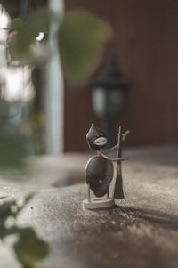 A whimsical, handmade sculpture crafted from stones and wire depicts a small humanoid figure holding a broom. The background is softly blurred, with warm tones suggesting an outdoor setting. A classic black outdoor lantern is faintly visible in the distance.