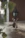 A whimsical, handmade sculpture crafted from stones and wire depicts a small humanoid figure holding a broom. The background is softly blurred, with warm tones suggesting an outdoor setting. A classic black outdoor lantern is faintly visible in the distance.