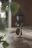 A whimsical, handmade sculpture crafted from stones and wire depicts a small humanoid figure holding a broom. The background is softly blurred, with warm tones suggesting an outdoor setting. A classic black outdoor lantern is faintly visible in the distance.