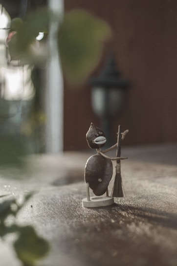 A whimsical, handmade sculpture crafted from stones and wire depicts a small humanoid figure holding a broom. The background is softly blurred, with warm tones suggesting an outdoor setting. A classic black outdoor lantern is faintly visible in the distance.