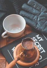 A brass cezve filled with freshly brewed coffee rests on a round wooden base, next to an empty white cup. A black book with partially visible white lettering lies beneath them. A dark, knitted fabric and a black, padded glove are nearby, suggesting a cozy setting.