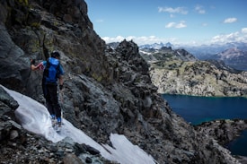 A person wearing hiking gear climbs a rocky and snowy mountain slope, using trekking poles for support. The scene includes a breathtaking view of a mountainous landscape with a large, blue lake and distant peaks under a partly cloudy sky.