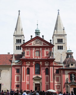 A vibrant, red and beige historic cathedral with two tall bell towers and a central ornate structure. The cathedral features intricate architectural details, including statues and relief carvings. Many people are gathered in front of the cathedral, adding to the sense of activity and liveliness.