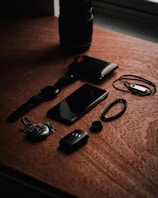 A set of personal items including a watch, smartphone, wallet, and various accessories are arranged on a wooden surface. The lighting creates a moody ambience with shadows cast across the objects.
