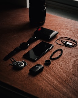A set of personal items including a watch, smartphone, wallet, and various accessories are arranged on a wooden surface. The lighting creates a moody ambience with shadows cast across the objects.