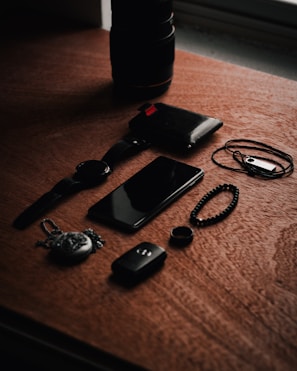 A set of personal items including a watch, smartphone, wallet, and various accessories are arranged on a wooden surface. The lighting creates a moody ambience with shadows cast across the objects.