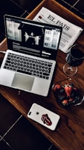 A stylish and cozy arrangement featuring a laptop with a webpage open, a Spanish newspaper titled 'El País', a glass of red wine, and a bowl of fresh strawberries and blackberries. The laptop and smartphone are placed on a wooden table, adding a warm and sophisticated touch.