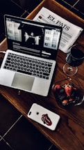 A stylish and cozy arrangement featuring a laptop with a webpage open, a Spanish newspaper titled 'El País', a glass of red wine, and a bowl of fresh strawberries and blackberries. The laptop and smartphone are placed on a wooden table, adding a warm and sophisticated touch.