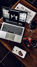 A stylish and cozy arrangement featuring a laptop with a webpage open, a Spanish newspaper titled 'El País', a glass of red wine, and a bowl of fresh strawberries and blackberries. The laptop and smartphone are placed on a wooden table, adding a warm and sophisticated touch.