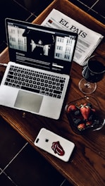 A stylish and cozy arrangement featuring a laptop with a webpage open, a Spanish newspaper titled 'El País', a glass of red wine, and a bowl of fresh strawberries and blackberries. The laptop and smartphone are placed on a wooden table, adding a warm and sophisticated touch.