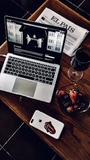 A stylish and cozy arrangement featuring a laptop with a webpage open, a Spanish newspaper titled 'El País', a glass of red wine, and a bowl of fresh strawberries and blackberries. The laptop and smartphone are placed on a wooden table, adding a warm and sophisticated touch.