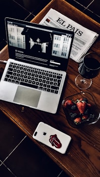 A stylish and cozy arrangement featuring a laptop with a webpage open, a Spanish newspaper titled 'El País', a glass of red wine, and a bowl of fresh strawberries and blackberries. The laptop and smartphone are placed on a wooden table, adding a warm and sophisticated touch.