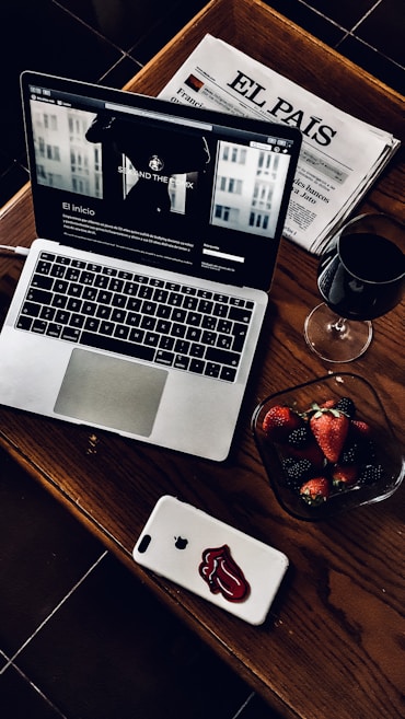 A stylish and cozy arrangement featuring a laptop with a webpage open, a Spanish newspaper titled 'El País', a glass of red wine, and a bowl of fresh strawberries and blackberries. The laptop and smartphone are placed on a wooden table, adding a warm and sophisticated touch.
