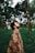 A person, wrapped in a patterned shawl, stands in the middle of a lush forest with their eyes closed and head tilted slightly upwards. Tall trees surround the individual, and a carpet of green foliage covers the ground, creating a serene and tranquil atmosphere.