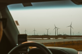 A view through a car windshield on a road with numerous wind turbines spread across a flat landscape. The scene includes a driver's silhouette, a mobile phone mount, and a vast expanse of sky above.