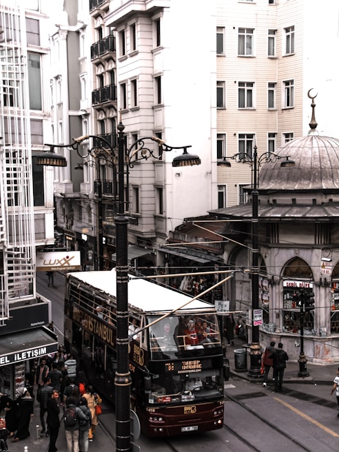 A bustling street scene in an urban area with several people walking on the sidewalk. A double-decker tour bus with 'Big Bus Istanbul' written on the side is seen driving down the street. The surrounding architecture features tall, modern buildings, and a few street lamps are visible. There is a small kiosk or shop with a decorative dome roof, and multiple signs and storefronts can be seen.