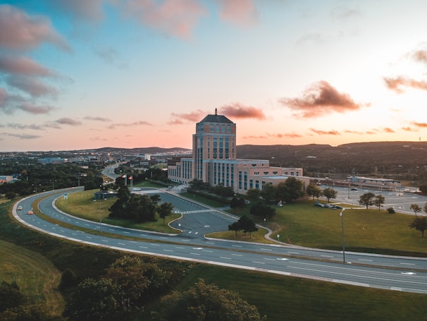 Aerial view of a large office or government building located near a highway interchange. The structure is surrounded by green lawns and trees, and there are cars visible on the road. The sky is partly cloudy with shades of orange and pink, indicating a sunrise or sunset.