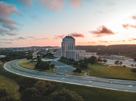 Aerial view of a large office or government building located near a highway interchange. The structure is surrounded by green lawns and trees, and there are cars visible on the road. The sky is partly cloudy with shades of orange and pink, indicating a sunrise or sunset.