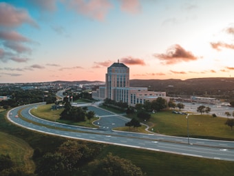 Aerial view of a large office or government building located near a highway interchange. The structure is surrounded by green lawns and trees, and there are cars visible on the road. The sky is partly cloudy with shades of orange and pink, indicating a sunrise or sunset.