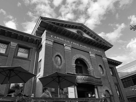 Historic building labeled as Carnegie Free Public Library with classic architectural features including brick facade, decorative arches, and large windows. Three people are visible in the foreground under open umbrellas, appearing to be conversing.