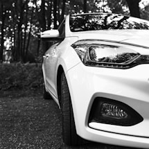 A close-up shot of the front portion of a white car, showing intricate details of the headlights and bumper, set against a backdrop of a wooded area, creating a reflective pattern on the car's surface.