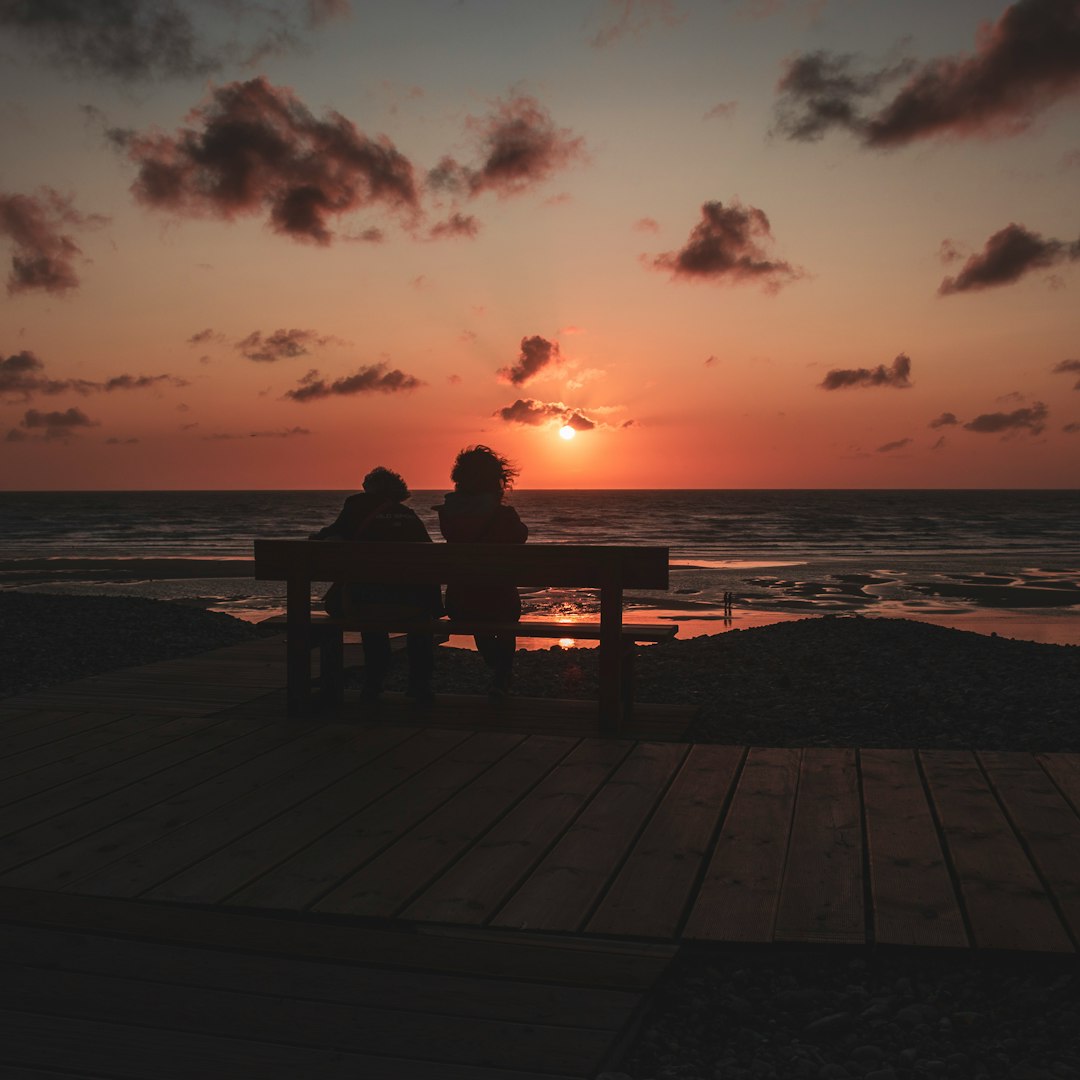 silhouette of man two person sitting on boardwalk bench looking at sunset seashore scenery
