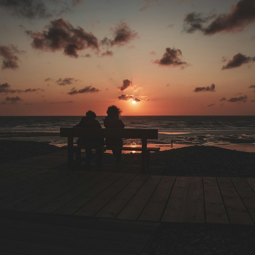 silhouette of man two person sitting on boardwalk bench looking at sunset seashore scenery