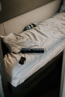A neatly made bed with a striped sheet and pillow is in a small, enclosed space. On the bed are a few electronic devices, possibly batteries or chargers, and two plastic bottles lying on the pillow.