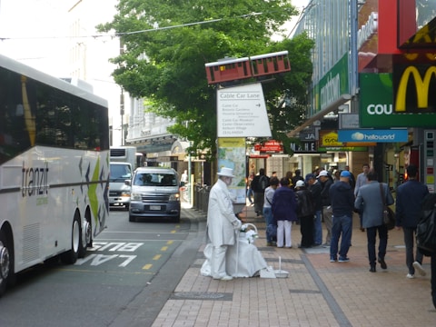 A busy street scene featuring a large white bus marked 'tranzit', with 'scenic tours' written below. People walk along the sidewalk, including a street performer dressed entirely in white, resembling a statue. Various signs and shopfronts, including a McDonald's, are visible. A small model of a red cable car is mounted above, adding a unique element to the urban setting.