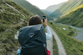A person with a backpack is capturing a scenic photograph on a trail surrounded by rolling hills and a winding river. The landscape is lush and expansive, indicating a remote and tranquil location. The sun casts a soft light, emphasizing the textures and contours of the hills.